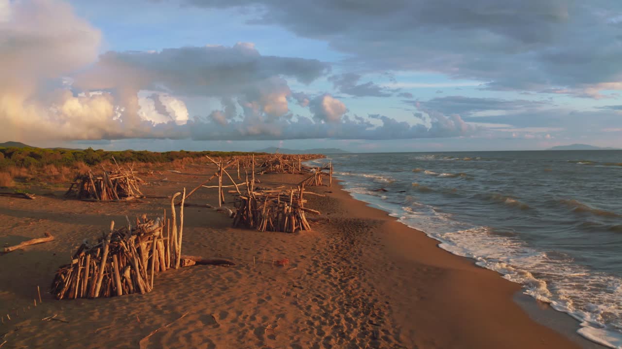 imágenes aéreas de drones de tipis de madera a la deriva en una playa de arena junto al mar en el icónico parque natural maremma en toscana, italia, con un espectacular cielo nuboso al atardecer con pequeñas olas azules y la isla giglio
