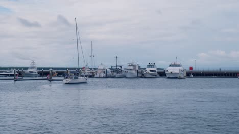 Un-Pequeño-Velero-Blanco-Ha-Entrado-En-El-Puerto-Deportivo-Y-Se-Está-Preparando-Para-Atracar,-Con-Otros-Barcos-Más-Grandes-Amarrados-Cerca