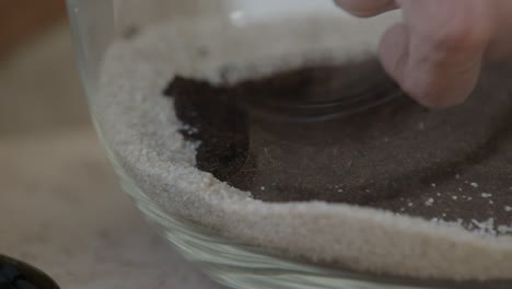 A-young-female-botanist-creates-a-tiny-live-forest-ecosystem-in-a-glass-terrarium---putting-soil-layer-on-sand---a-tight-close-up