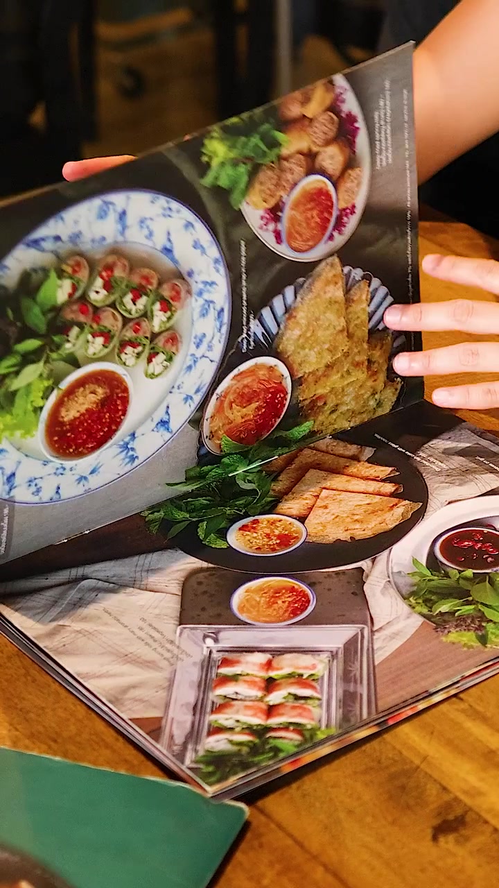 Hænder bladrer igennem en vietnamesisk madmenu på et træbord i et varmt oplyst restaurantmiljø.