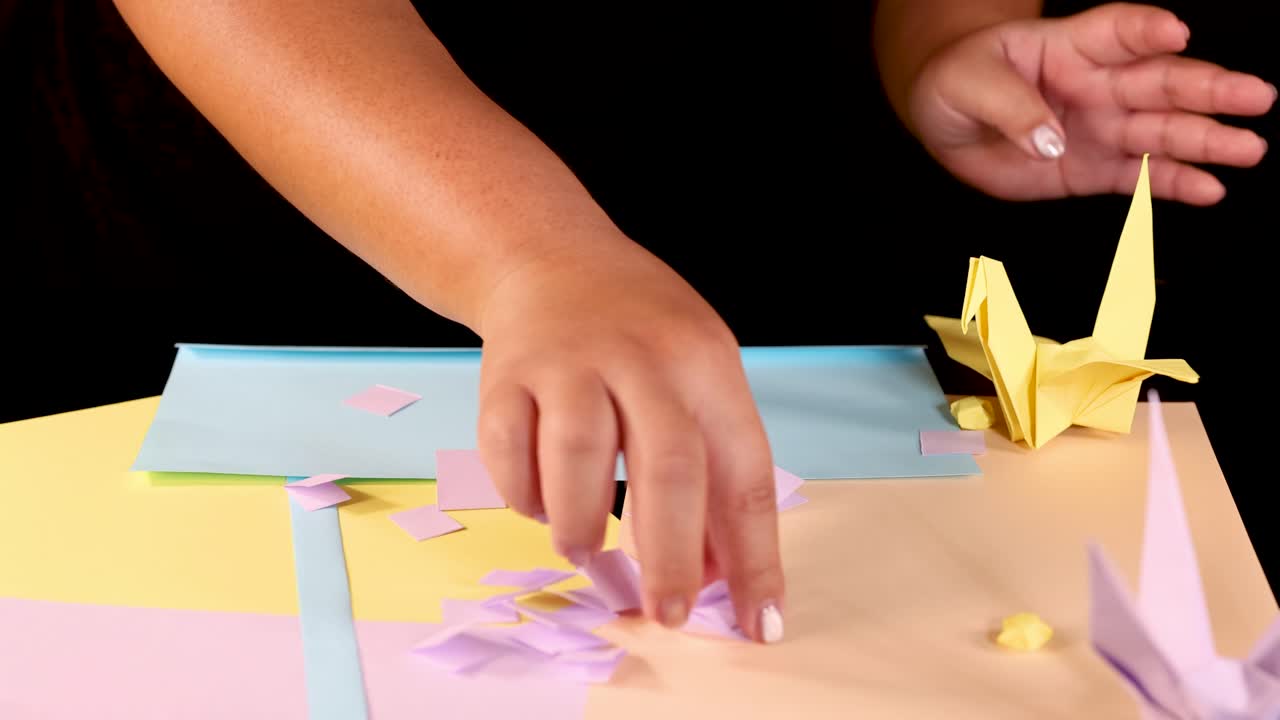 Person arranges yellow origami crane and pastel paper squares on brightly lit craft table