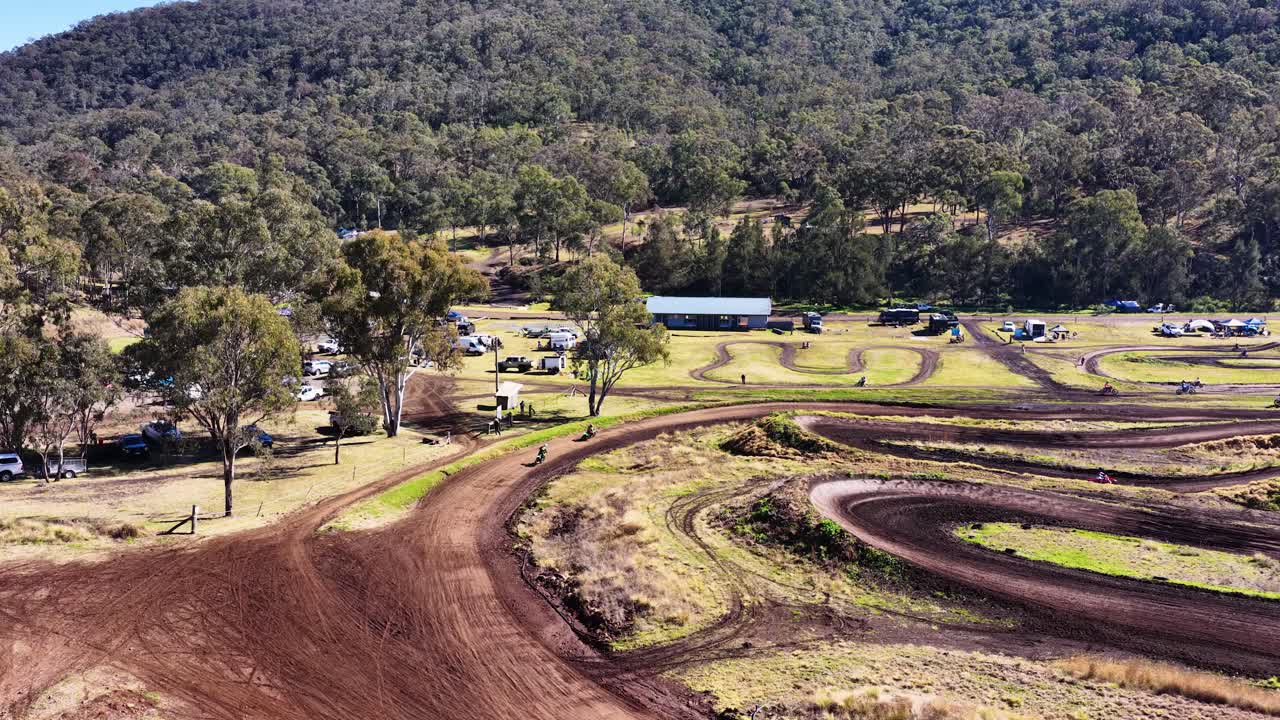 Drone footage glides over winding motocross track, sunlit landscape, trees, and distant hills