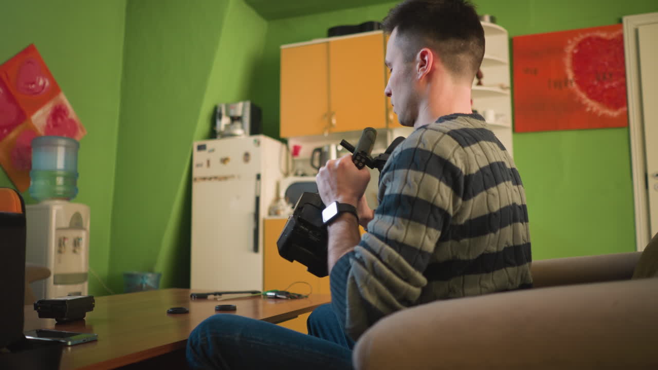 Person seated, adjusting video camera, attaching microphone, preparing filming equipment. Indoor workspace with tech tools, camera gear, and accessories, professional production setup