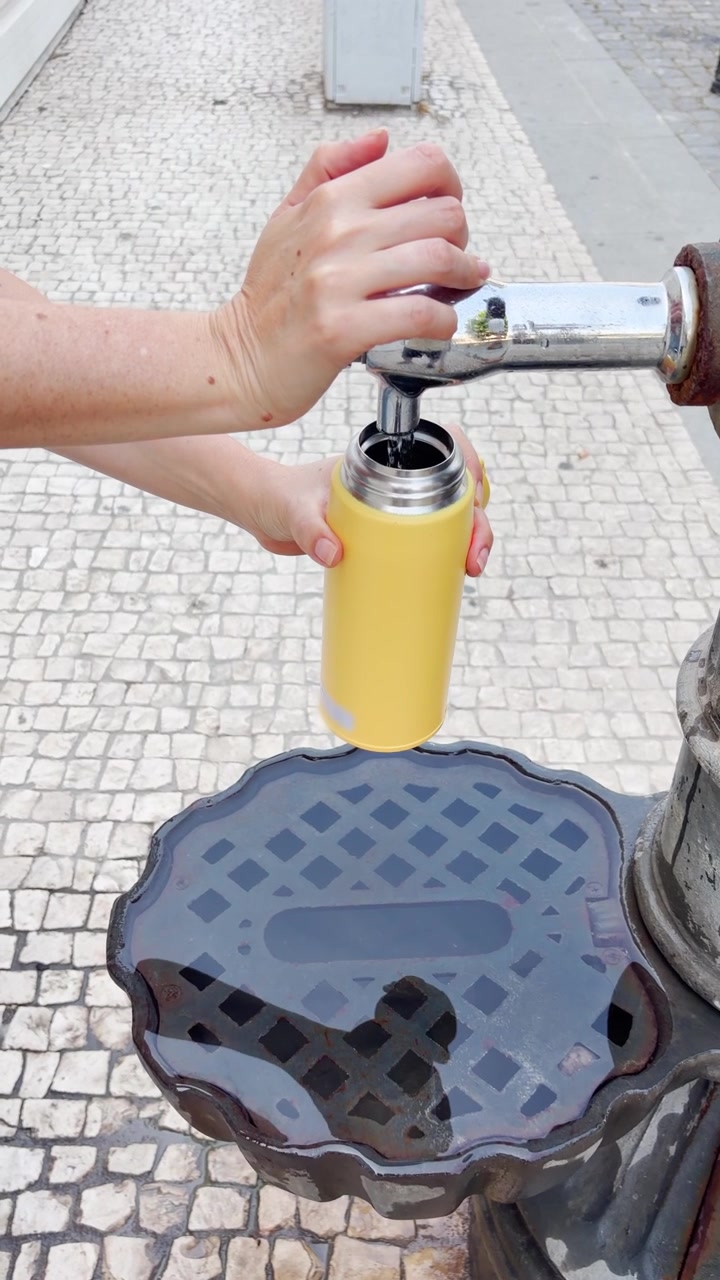 Donna che riempie una bottiglia d'acqua da un rubinetto esterno in un paese europeo durante un viaggio. Giornata calda all'aperto, con una turista o una persona del posto che usa una fontana pubblica di acqua potabile. Strada di Porto, Portogallo. Reel in stile UGC.