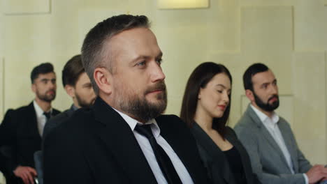 caucasian mature businessman sitting on a chair among people in a conference room, then he turns his face to the camera and smiling