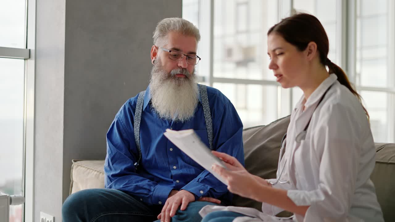 A brunette woman in a white medical coat tells an elderly man with gray hair and a lush beard in a blue shirt about his need to improve his health during a home examination on the sofa in a modern apartment