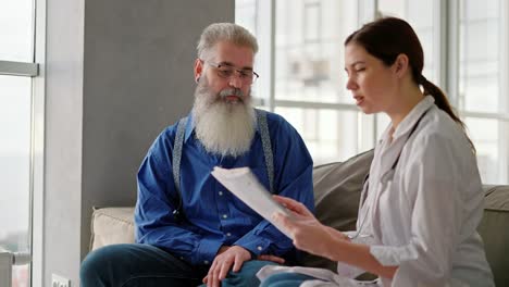 a brunette woman in a white medical coat tells an elderly man with gray hair and a lush beard in a blue shirt about his need to improve his health during a home examination on the sofa in a modern apartment