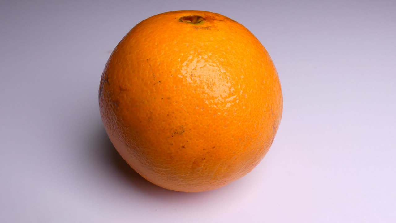 An orange in a white background. The light source slowly moves above the fruit slightly changing the shadows.
