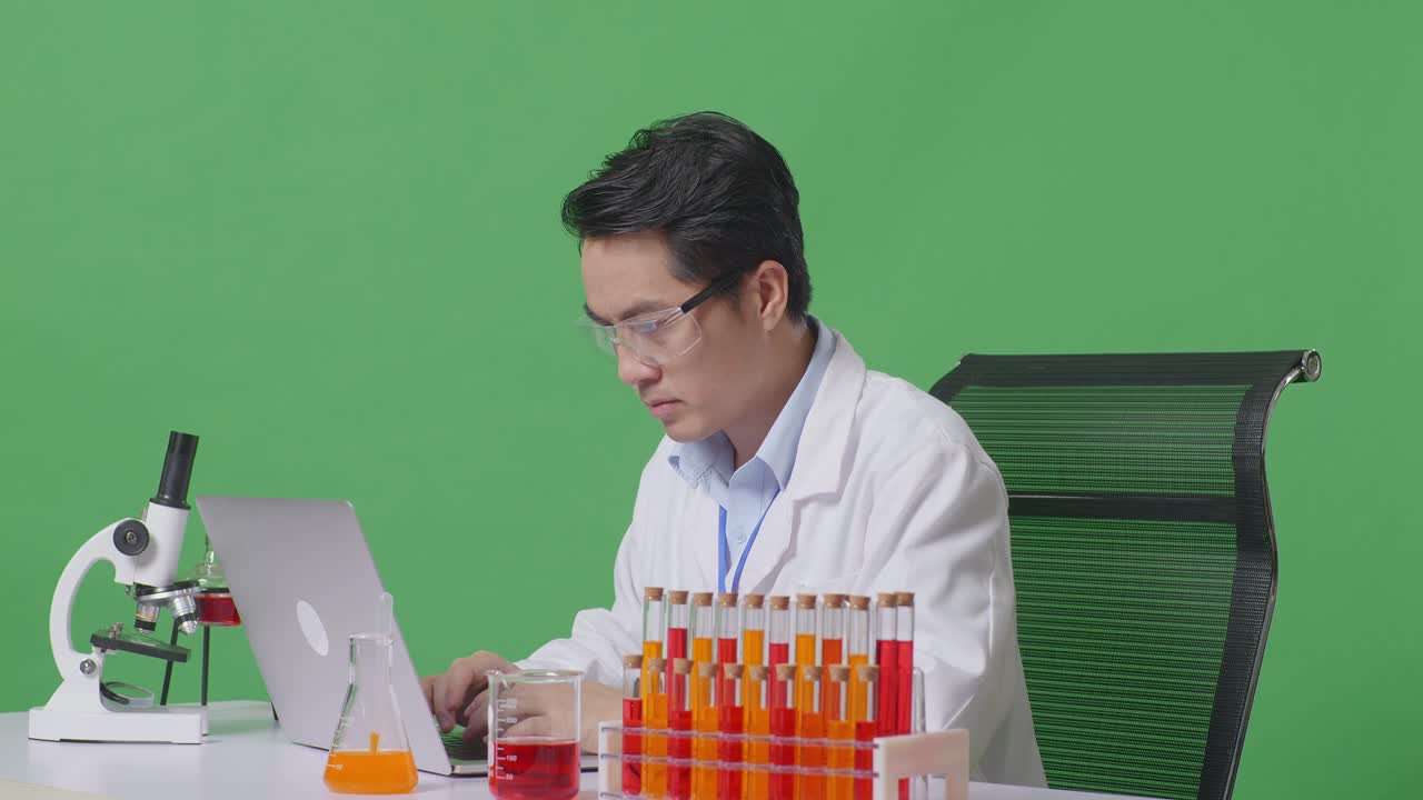 Side View Of Asian Man Scientist Typing On A Laptop While Working On The Table With Microscope And Test Tube In The Green Screen Background Laboratory