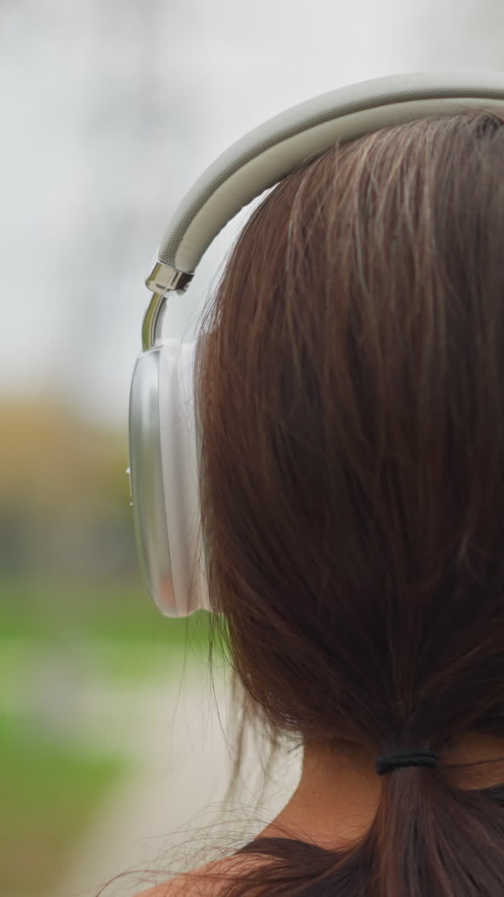 Vista posteriore di una giovane donna che si mette le cuffie sulla testa, sentendosi rilassata mentre si gode la musica all'aperto in un ambiente naturale e calmo con una messa a fuoco morbida su di lei e sullo sfondo.