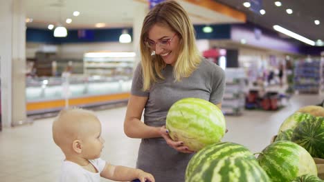 Young-mother-is-choosing-a-watermelon,-showing-it-to-her-little-child-which-is-sitting-in-a-grocery-cart.-Family-shopping-in