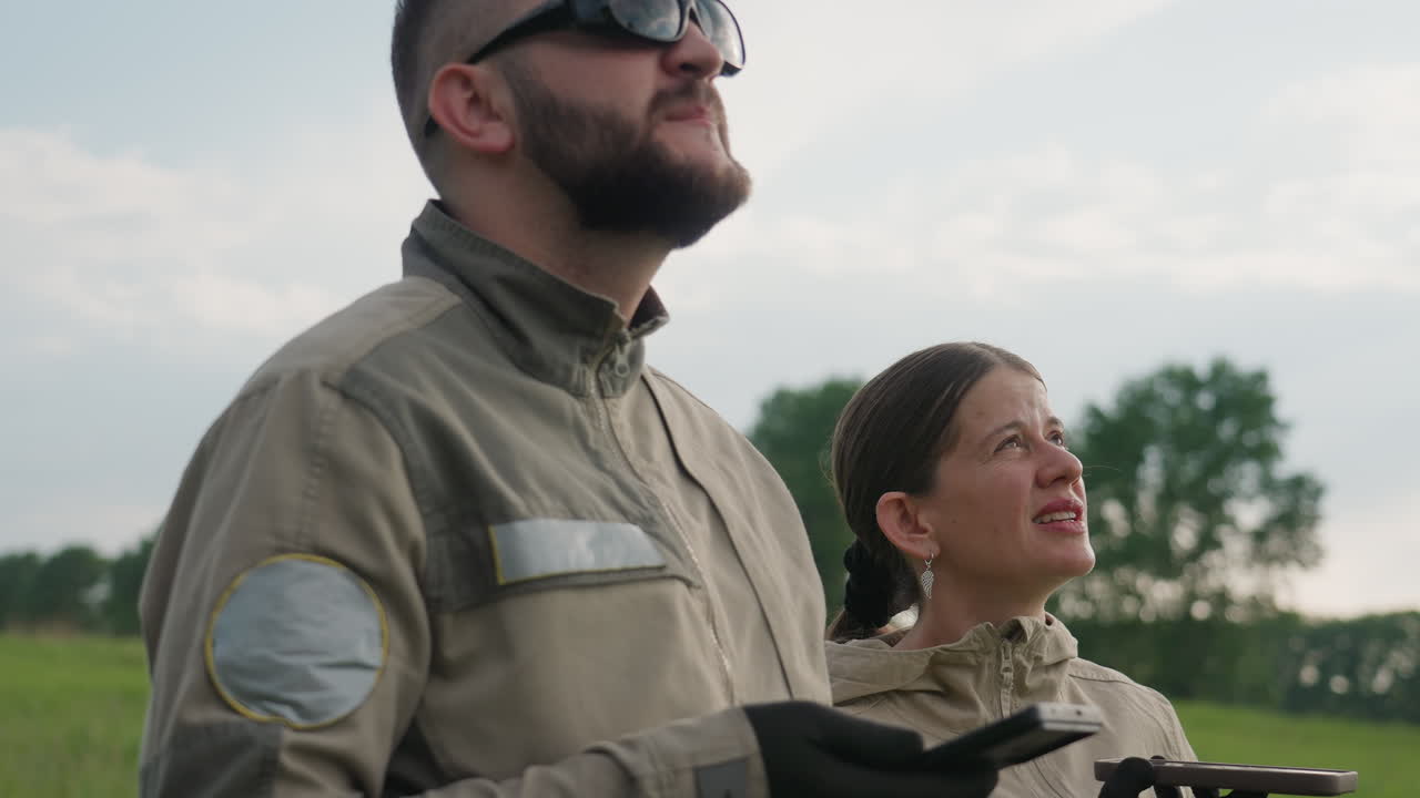 two colleagues stand in lush green field operating drone controllers and inspecting aerial device through handheld screens while insects circle overhead