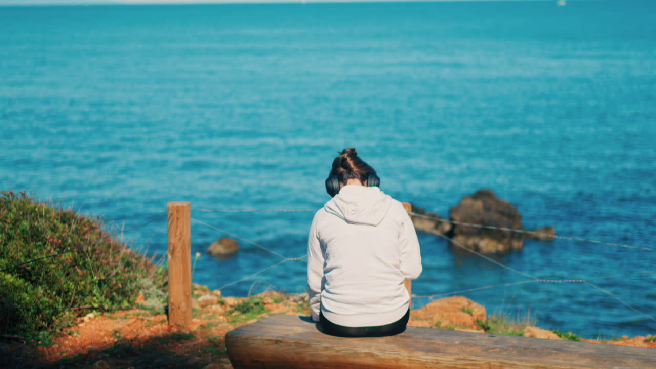 Woman with headphones on her head sitting on a wooden bench with a view of the sea