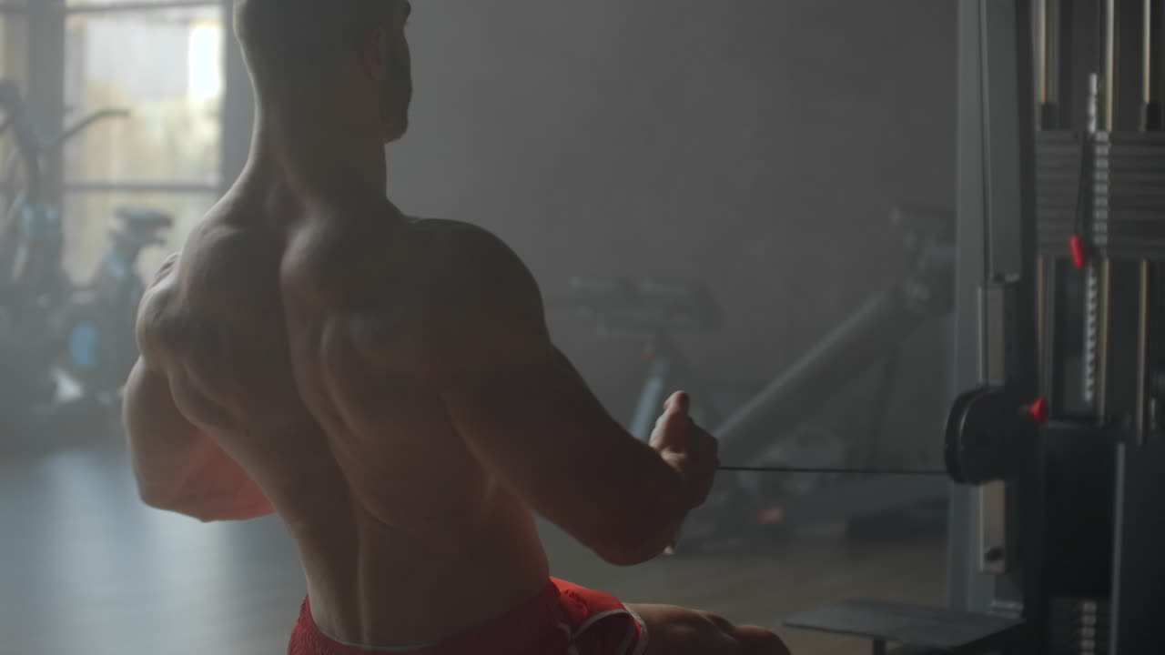 A man pulls weight to his stomach sitting in the simulator performing an exercise for the muscles of the back against the background of large Windows of the gym. Powerful male athlete doing rowing exercises