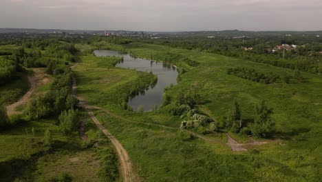 Vista-Aérea-De-Un-Lago-Rodeado-De-Exuberante-Vegetación,-Con-Senderos-Y-Edificios-Distantes