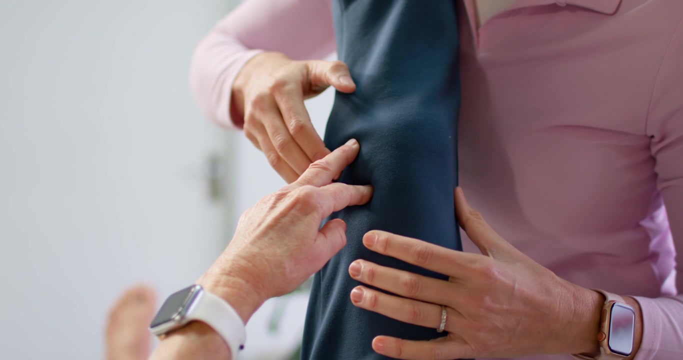 Two adult females holding raised leg while one palpating dark pants fabric during fitting check