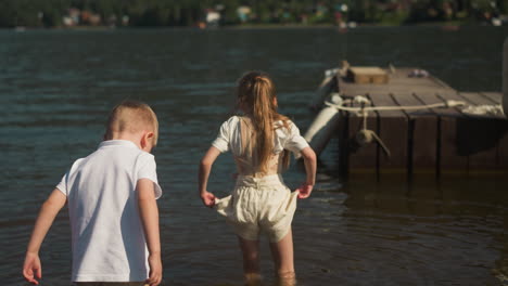 girl in corset to correct posture walks knee deep in water ahead of boy. adventures for children in resort area. daughter carefully holds edges of shorts