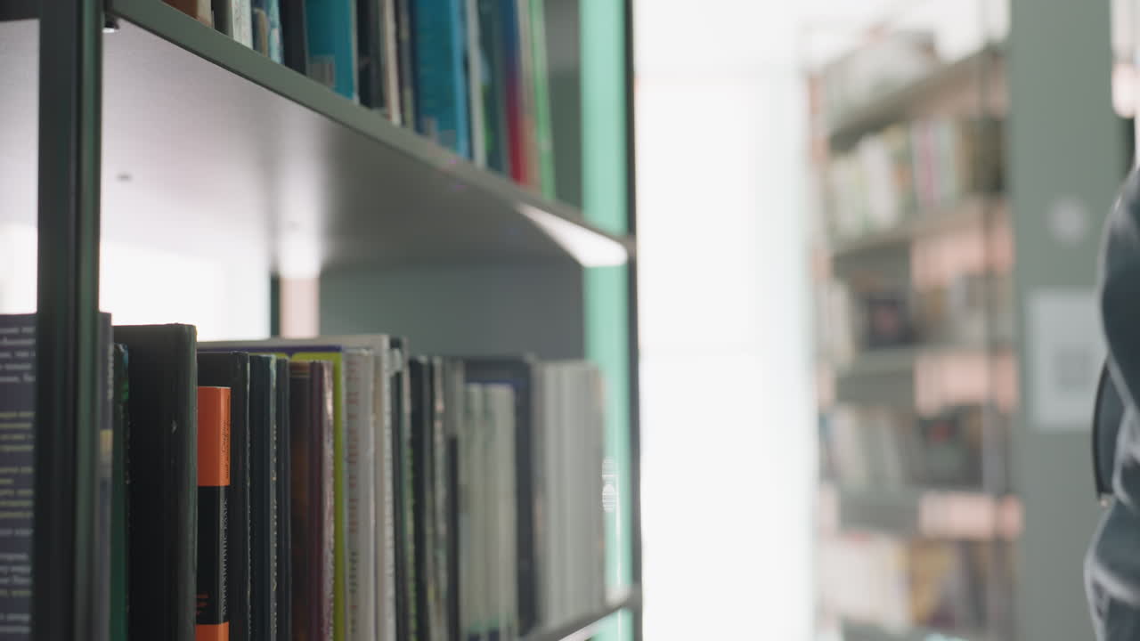 Close up of focused student in casual clothing returning book to shelf in bright library, surrounded by rows of books under soft daylight, creating studious and calm atmosphere of reading space