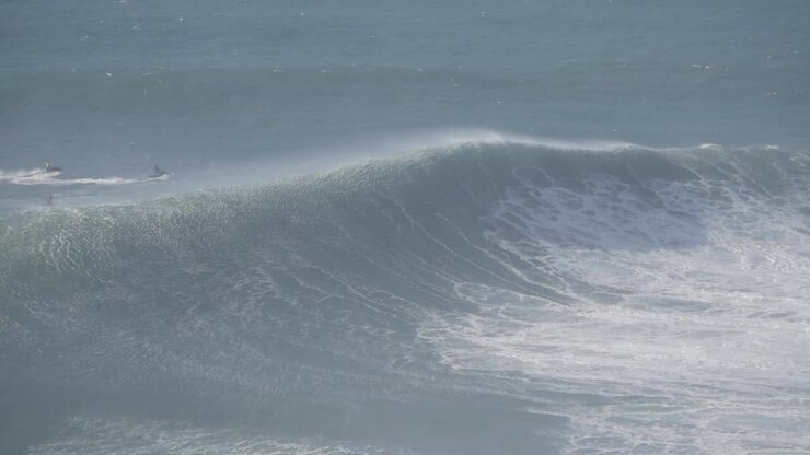 Massive wave rolling at Nazaré, Portugal, with surfers visible in the distance