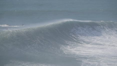 Massive wave rolling at Nazaré, Portugal, with surfers visible in the distance