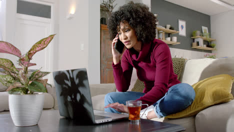 happy biracial woman using laptop and talking on smartphone at home, slow motion