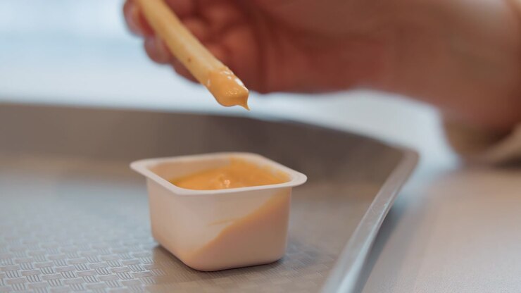 Una mano tiene un bastoncino di legno per mescolare ricoperto da una piccola quantità di salsa cremosa all'arancia sopra una piccola tazza quadrata di plastica per condimenti poggiata su un vassoio di metallo testurizzato; movimento visibile e una leggera sfocatura sulla mano e sul bastoncino.