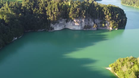 Die-Abendsonne-Wirft-Lange-Schatten-über-Den-Schweizer-Lac-De-Greyerz-Mit-Einem-Imposanten-Felsen-Im-Vordergrund-Und-Einem-Kleinen-Dorf-Im-Hintergrund,-Sonniges-Wetter