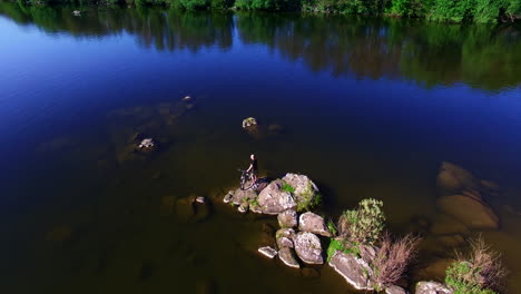 dark blue river with calm smooth surface. shallow water with stone peeping out of it. man standing on the rock with bicycle. top view.