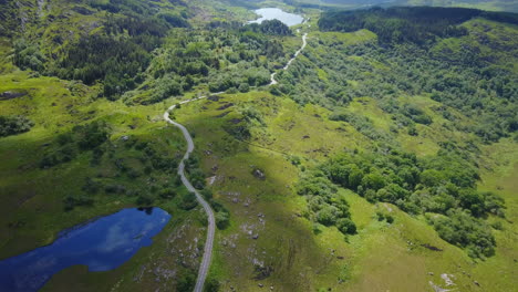 Amazing-high-aerial-video-of-reflective-blue-lake-in-foreground-with-long-winding-road,shadow-of-moving-clouds-passing,motor-bikers-in-distance-and-other-lakes-and-mountain-range-in-background