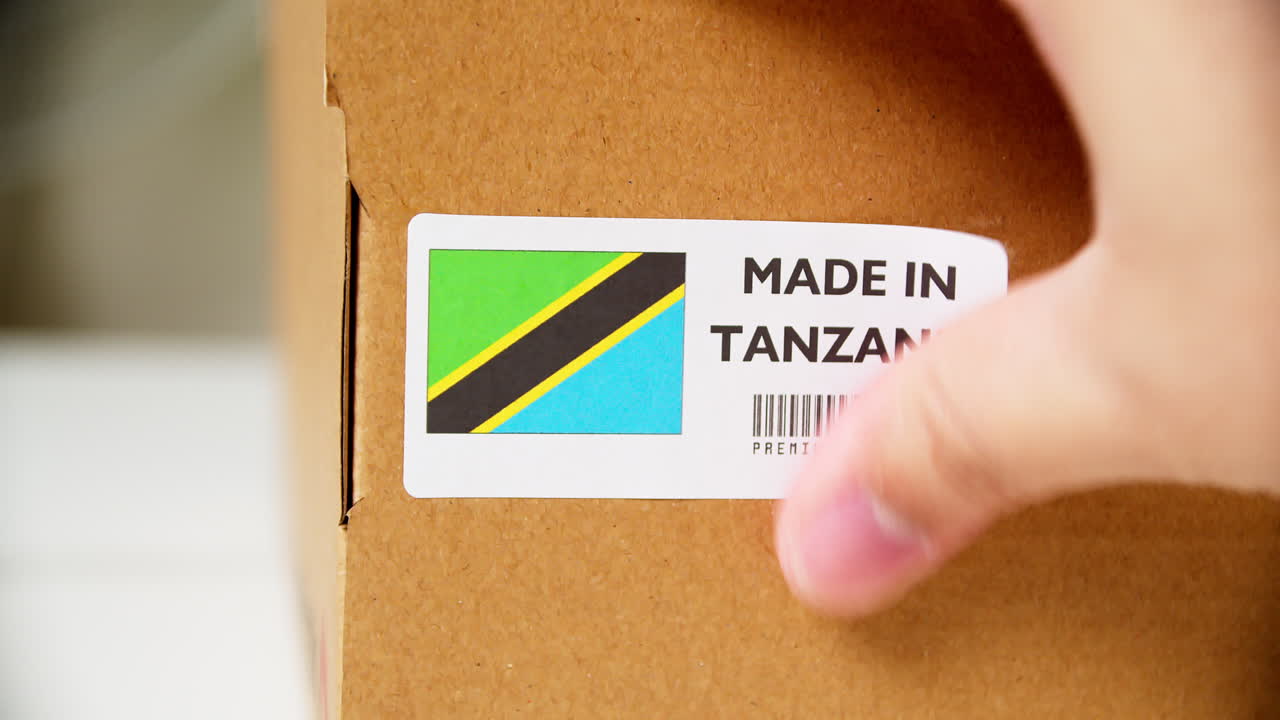 manos aplicando la etiqueta de la bandera de tanzania en una caja de cartón de envío con productos