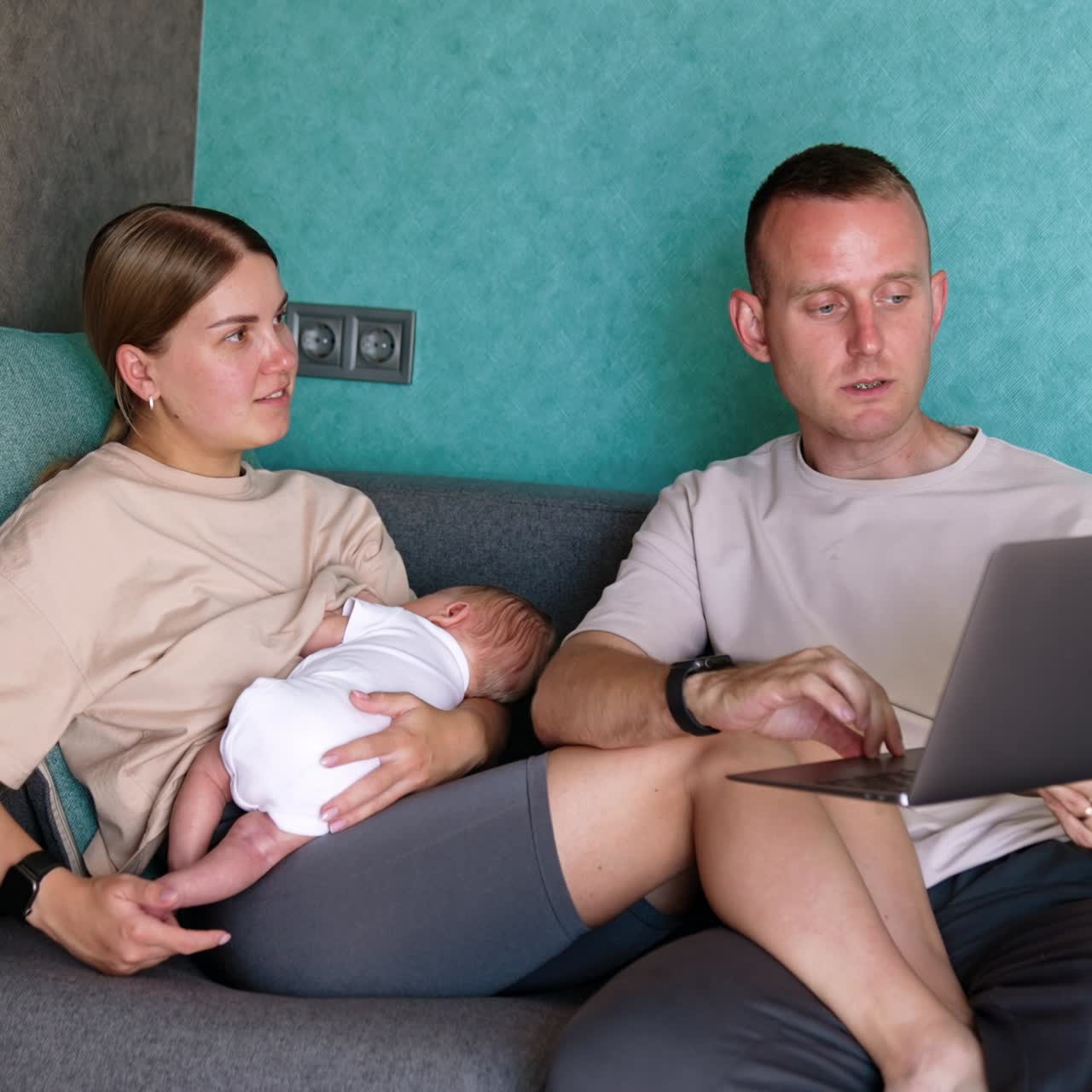 Caucasian family are in the room sitting on sofa. Parents of a newborn child looking at laptop in male arms