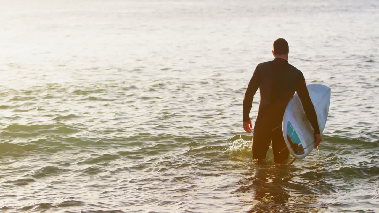vista trasera de un surfista caucásico adulto con tabla de surf caminando en el mar durante la puesta de sol 4k