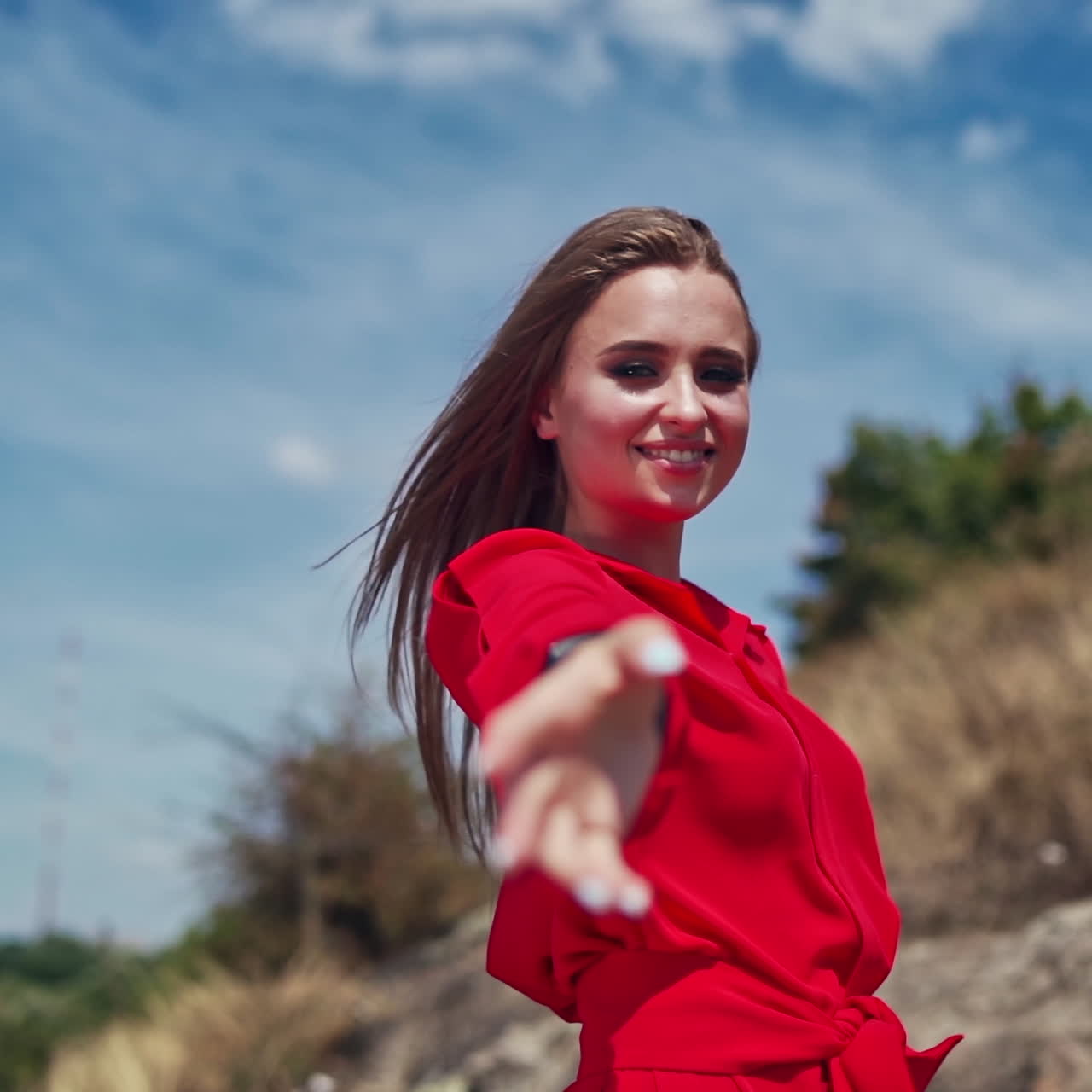 Sexy lady outdoors. Portrait of a young woman in red dress looking on camera and smiling on the natural background. Beautiful female attracts attention by her hand.