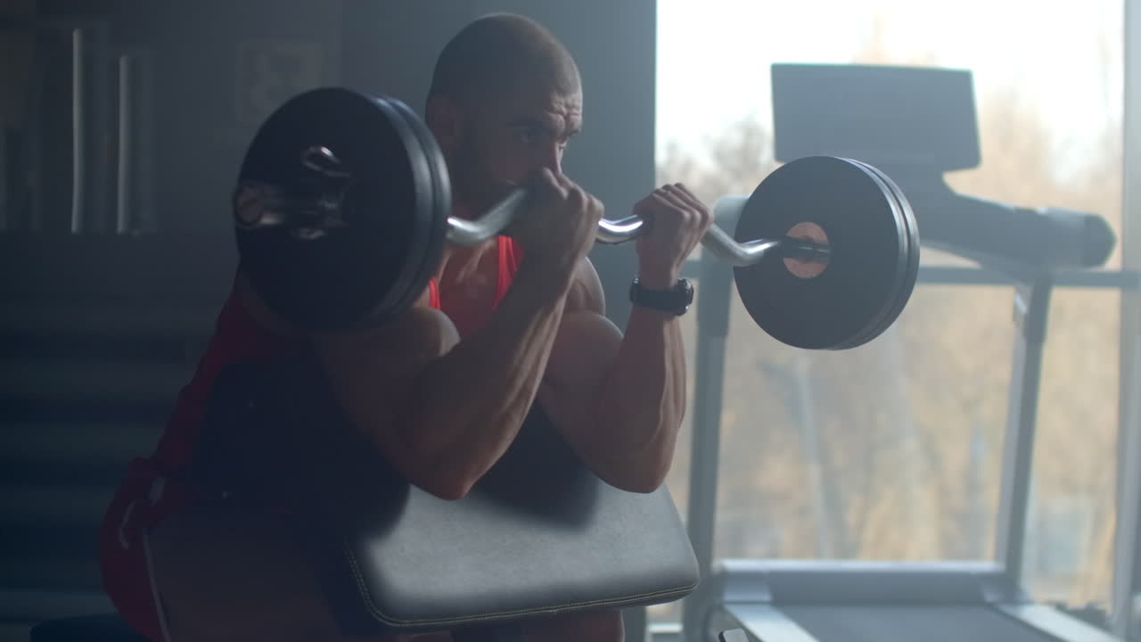 A strong male bodybuilder lifts a weight barbell while sitting on a bench and trains hard in slow motion at the GYM. A bodybuilder trains professionally. Powerful strong hands