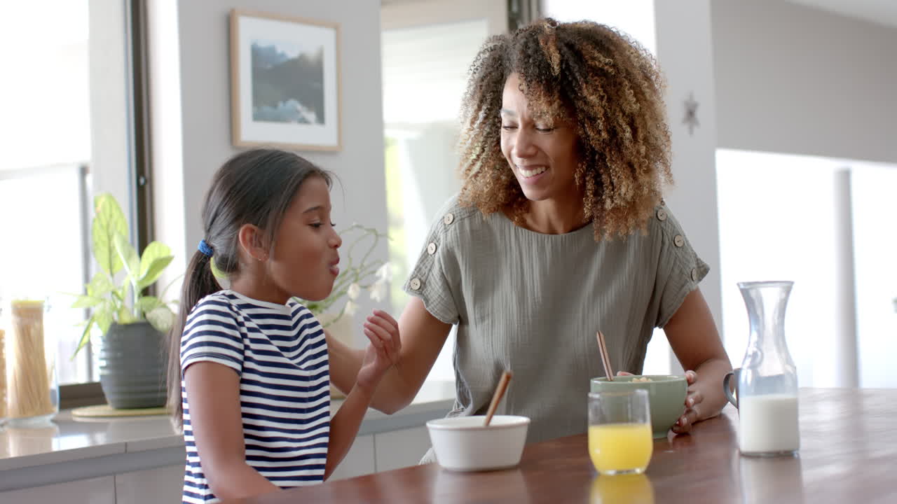 madre y hija bi-raciales felices comiendo cereales para el desayuno en una cocina soleada, cámara lenta