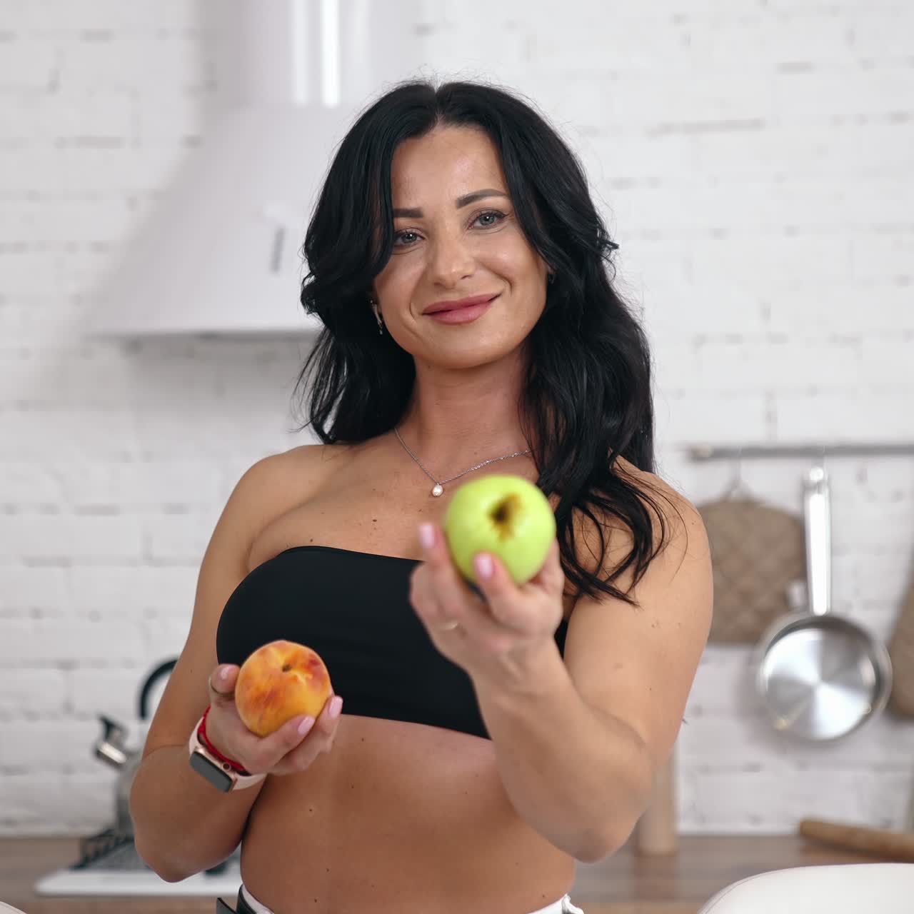 Attractive female proposes fresh apples indoors. Portrait of a woman with bare shoulders and belly holding juicy fruit and showing on camera in the kitchen