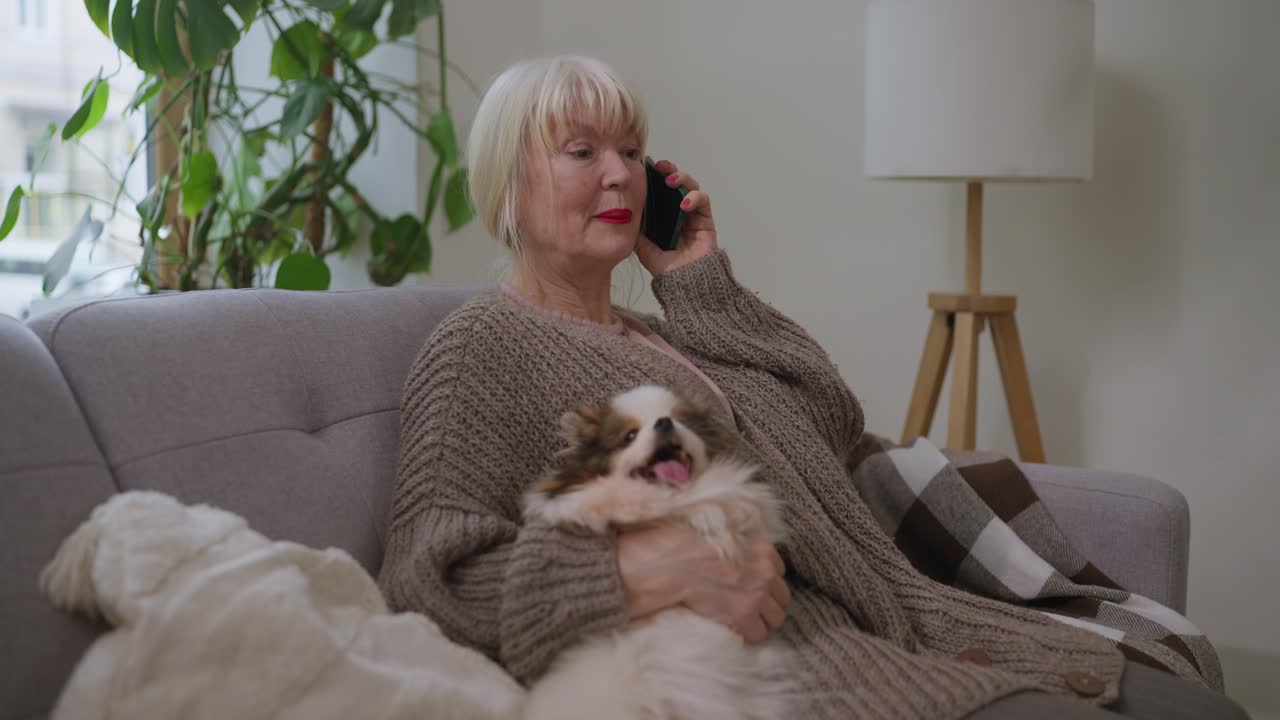 A senior woman is talking on her phone with a dog on her lap