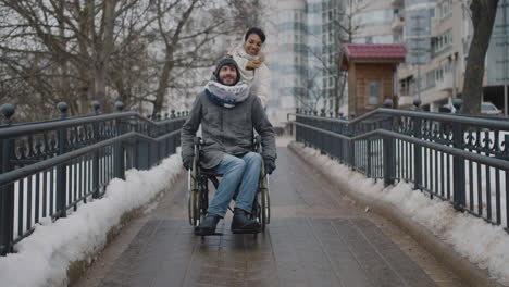 Front-View-Of-A-Muslim-Woman-Taking-Her-Disabled-Friend-In-Wheelchair-On-A-Walk-In-City-In-Winter
