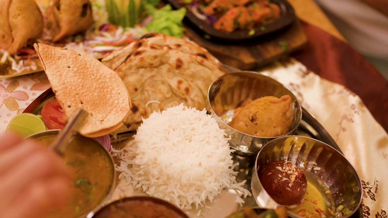 Close-up shot of hand placing papadum on a diverse Indian meal platter with warm lighting.