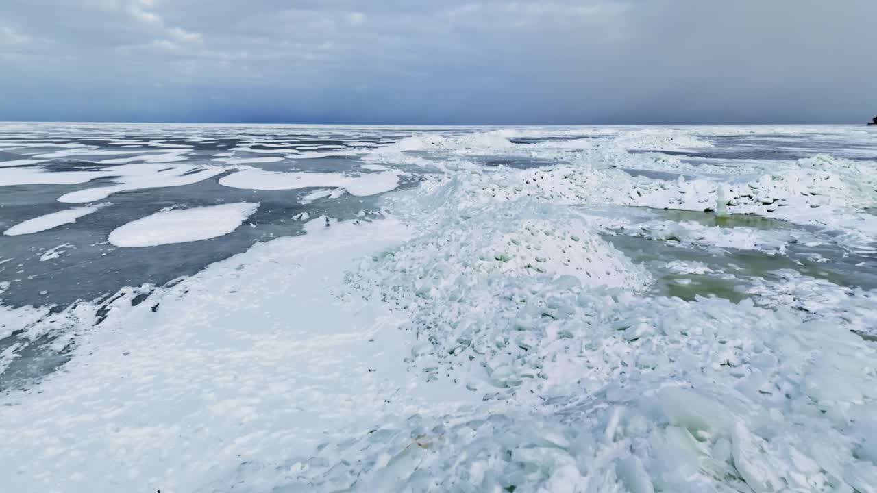 Sorvolo con drone di una costa ghiacciata con torri di ghiaccio accumulate, pozze di fusione fangose e enormi creste di pressione sotto un drammatico cielo invernale.