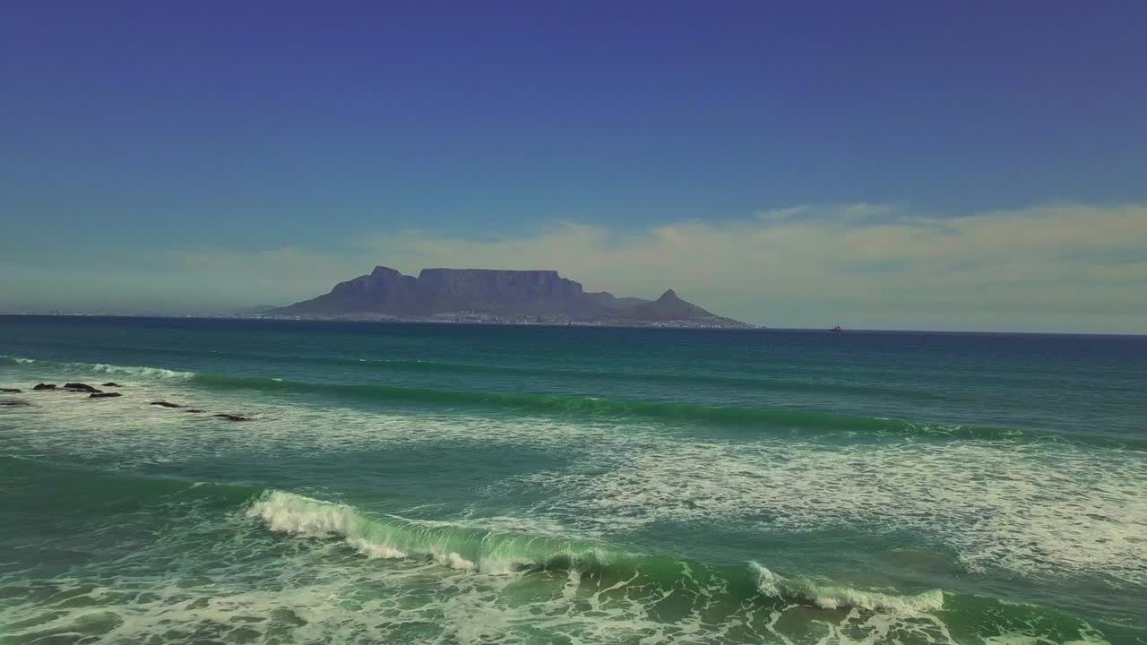 Aerial shot of Cape Town’s Table Mountain from across the Bay. The camera pulls back toward the beach in a cinematic pull back seeing the beautiful Atlantic shoreline and Table Mountain