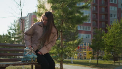 una joven con una chaqueta de melocotón, camisa interior gris y pantalones negros camina por un parque iluminado por el sol, se sienta en un banco y coloca sus patines a su lado