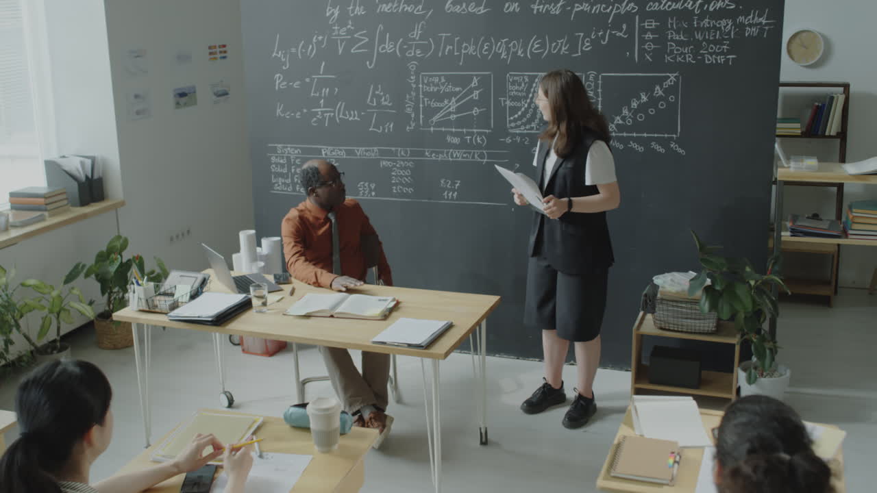 A professor lectures to students in a classroom with equations and charts on a blackboard