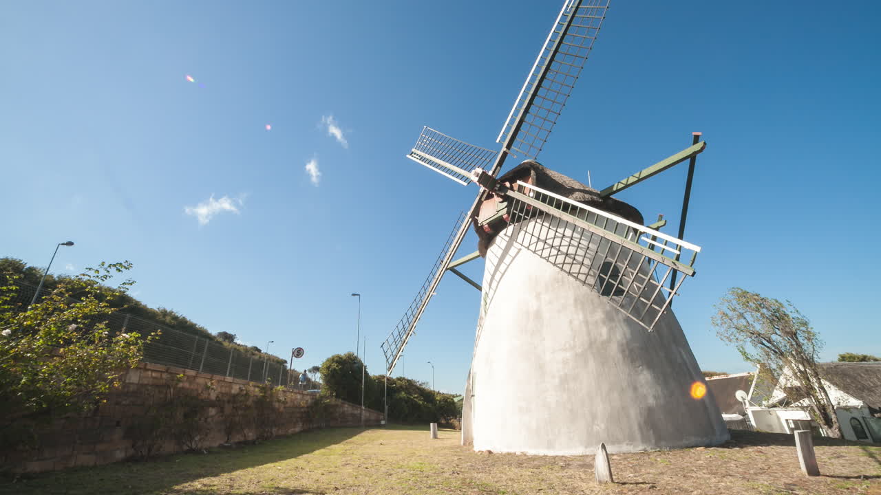 A motion control time-lapse at a classic windmill, which stands tall against a clear blue sky, blades ready to catch the wind. The iconic structure is framed by the bright day and lush greenery