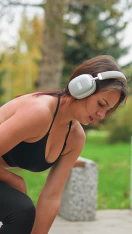 pretty athlete squatting left and right in outdoor park setting near concrete bench, wearing black activewear and listening to music with headphones, focusing on her workout