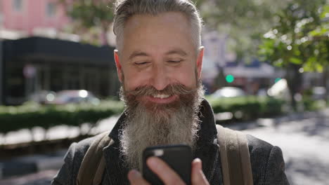 Retrato-De-Un-Hombre-Barbudo-Maduro-Y-Sofisticado-Sonriendo-Feliz-Usando-Mensajes-De-Texto-En-Un-Teléfono-Inteligente-Navegando-En-Una-Concurrida-Calle-De-La-Ciudad