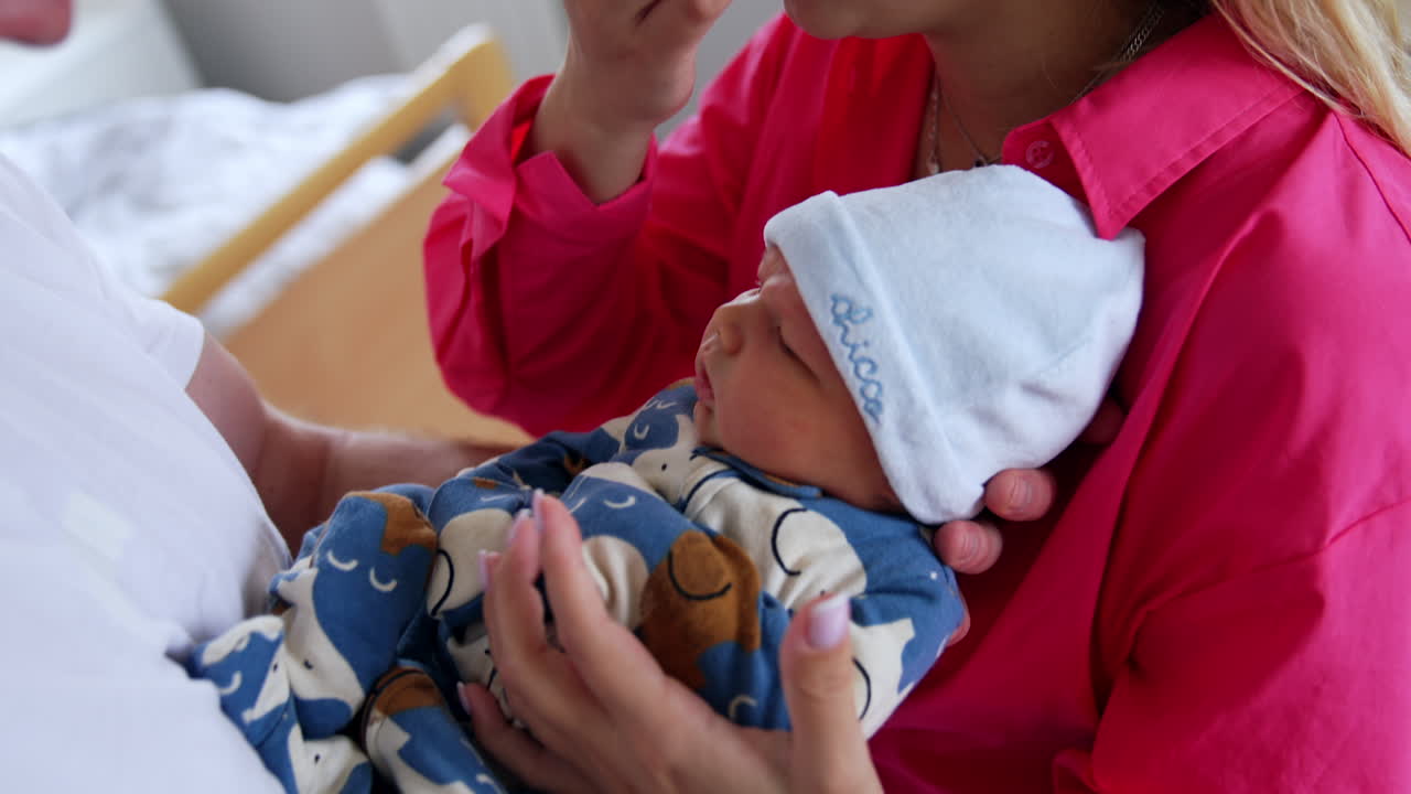 Tiny newborn in romper and cap lies between his parents on their hands. Mom touches baby nose with finger tenderly. Close up.