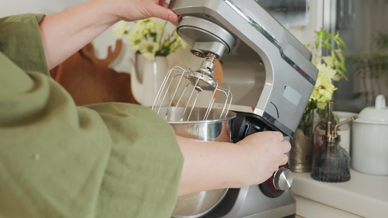 Student opens electric stand mixer to remove mixing attachment and stainless steel bowl filled with flour mixture in bright kitchen setting, demonstrating baking preparation process