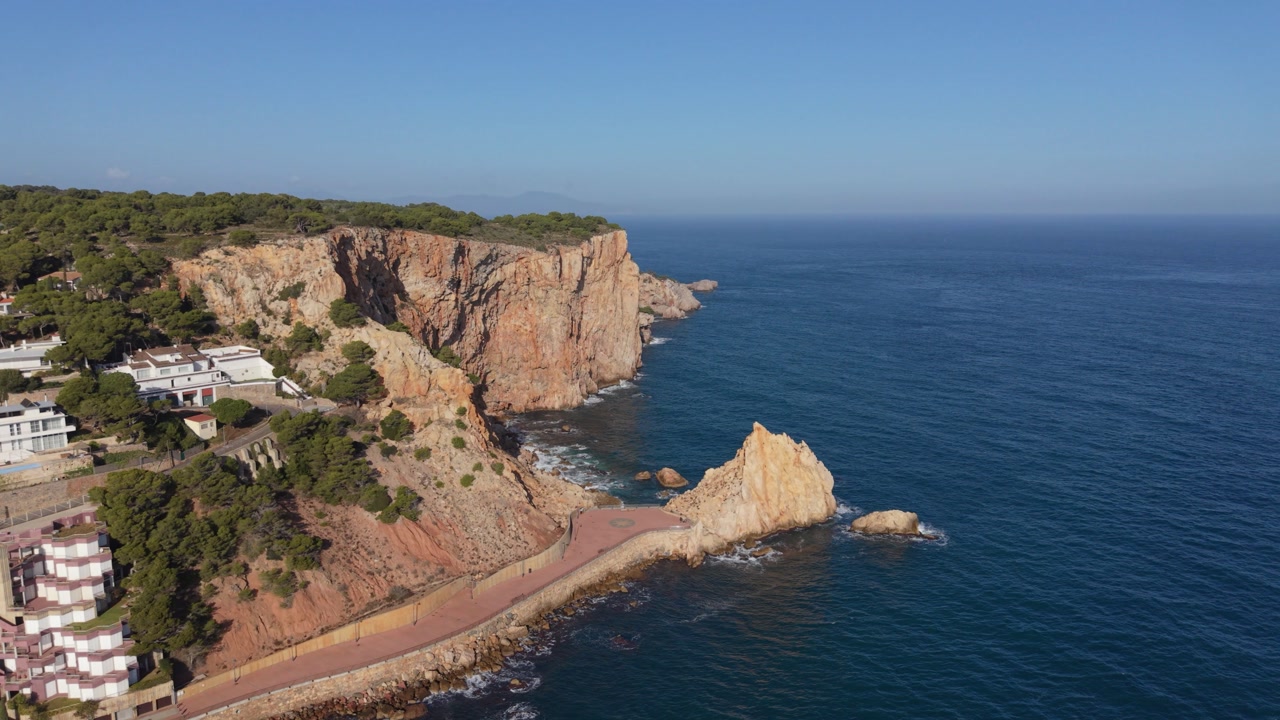 L’Estartit,Costa Brava,Spain high reveal drone footage of majestic cliffs rocky shoreline and the edge of town with cliff top and seaside properties in golden sun with clear blue sky and deep blue sea
