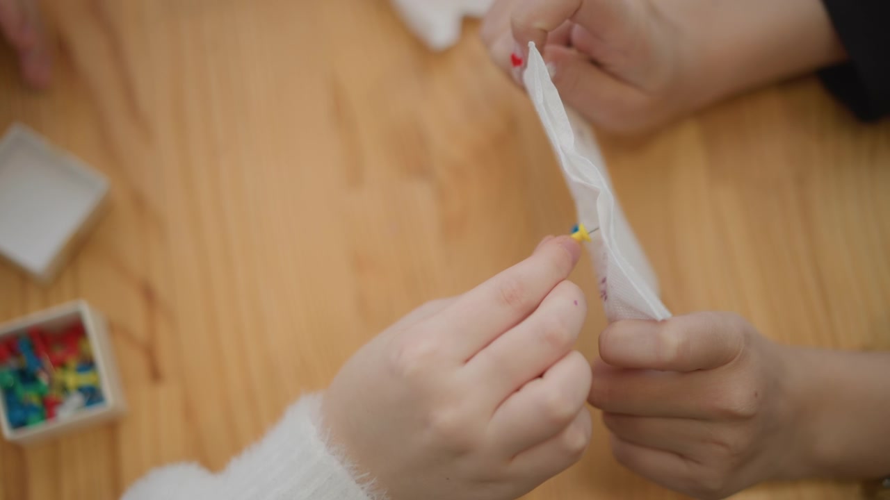 Primer plano cenital de manos de adulto y niño sobre una cálida mesa de madera, mientras el adulto abre un sobre blanco y el niño pellizca una pequeña pieza amarilla para recibirla, mostrando una guía cuidadosa e interacción compartida.